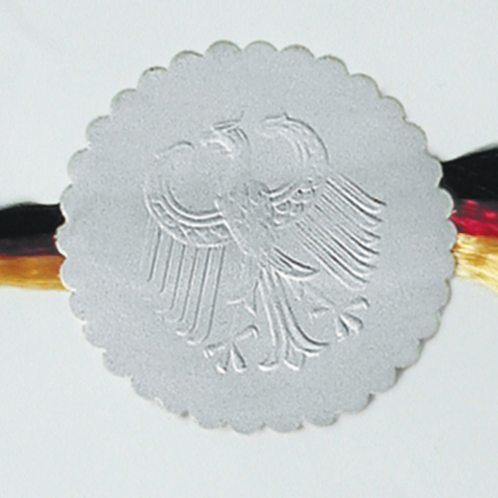 Geprägte Siegelmarke mit Adlerrelief Beispiel Beispiel einer geprägten Siegelmarke aus Papier mit Adlerrelief und gewelltem Rand, dahinter ein Band in Schwarz, Rot und Gold.