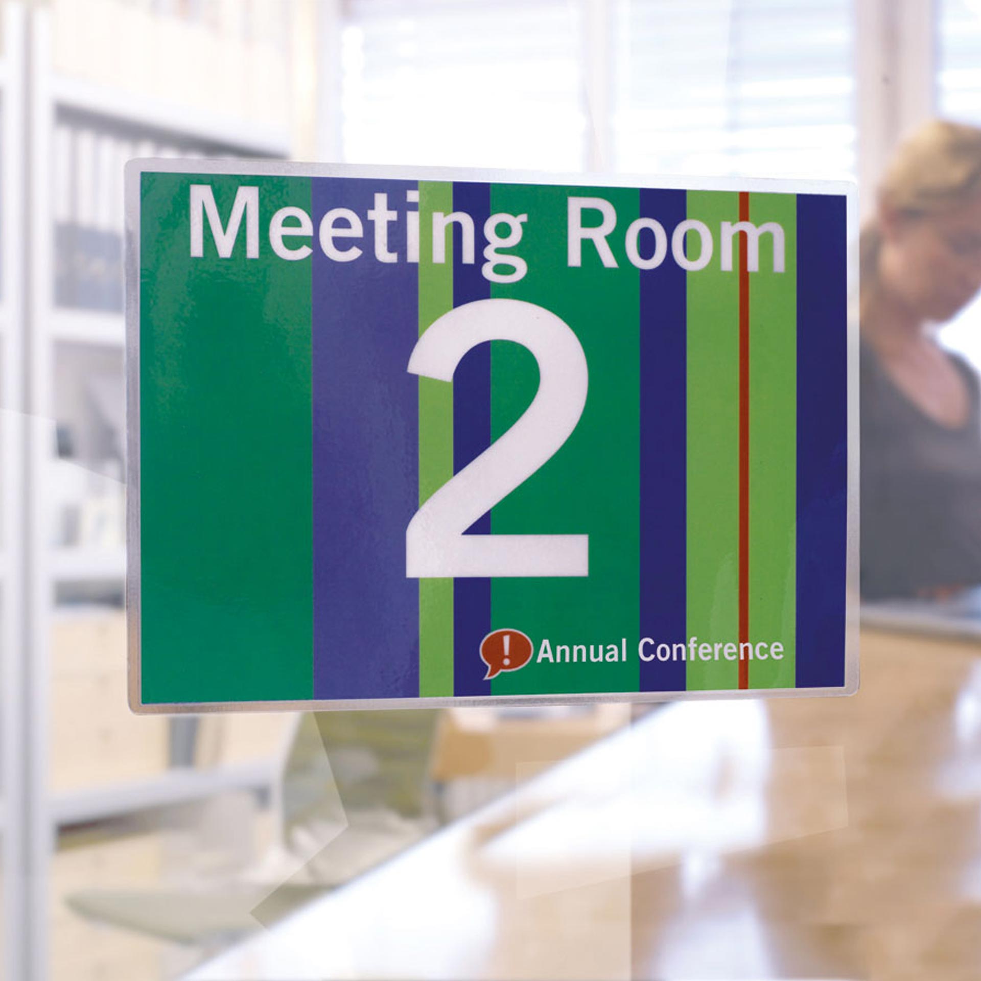 Laminiertes Schild „Meeting Room 2“ an einer Glaswand Laminiertes Schild an einer Glaswand mit der Aufschrift „Meeting Room 2“ und „Annual Conference“. Im unscharfen Hintergrund ist eine Person in einem Büroraum zu sehen.