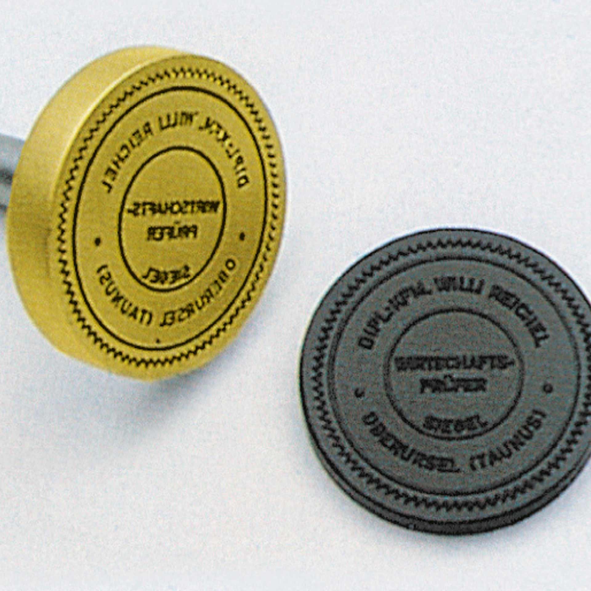 PERNUMA Prägestempel Beispielbild Zwei runde Prägestempel auf hellem Hintergrund, ein goldfarbener Oberstempel aus Metall und ein grauer Unterstempel mit kreisförmiger Beschriftung.