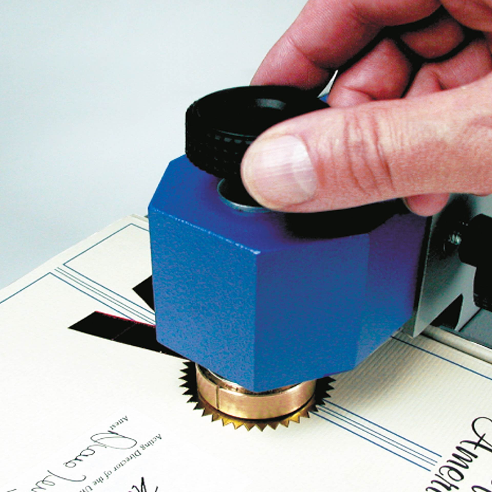 PERNUMA EMBOSSET Prägemaschine Detailansicht Stempelkopf Detailansicht der PERNUMA EMBOSSET Prägemaschine während der Nutzung. Eine Hand dreht den oberen Einstellknopf am blauen Stempelkopf, während ein Dokument unter dem Prägestempel liegt.